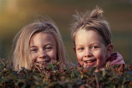 Bilde av ansiktet til to blide barn som dukker opp bak en hekk. Gutt og jente med blondt hår.  - Klikk for stort bilde